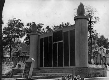 Emlékmű névtáblákkal egy temetőben Emlékmű névtáblákkal, két oszlop között, temetőben sírkövekkel a háttérben.