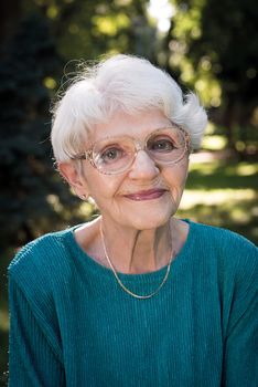 Idős nő kék ruhában arany nyaklánccal Idős nő kék ruhában arany nyaklánccal, zöld növényzet háttérrel